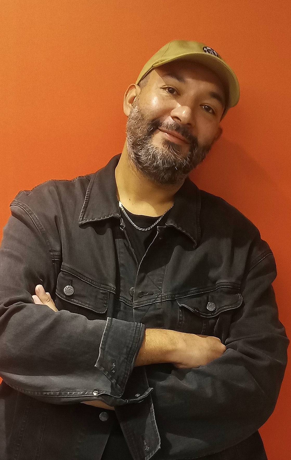 a man with a beard and a black jacket stands in front of an orange wall.